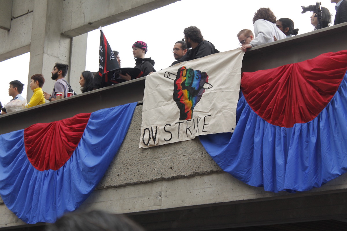A group of protesters stand on a balcony at Simon Fraser University. They have hung a banner that says ‘On Strike.’