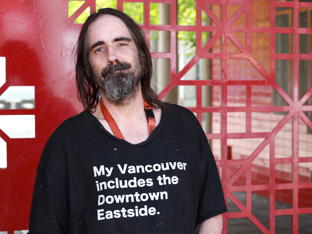 A man with shoulder-length salt-and-pepper hair and a beard looks at the camera. He&rsquo;s wearing a black T-shirt that reads, &lsquo;My Vancouver includes the Downtown Eastside.&rsquo;