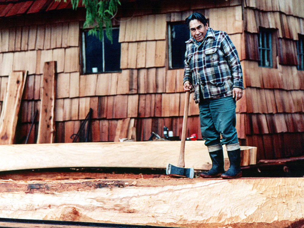 A man stands in front of a cedar cabin wearing a blue plaid jacket and holding a large axe. He&rsquo;s standing on a log which is being carved into a canoe using a single piece of wood.