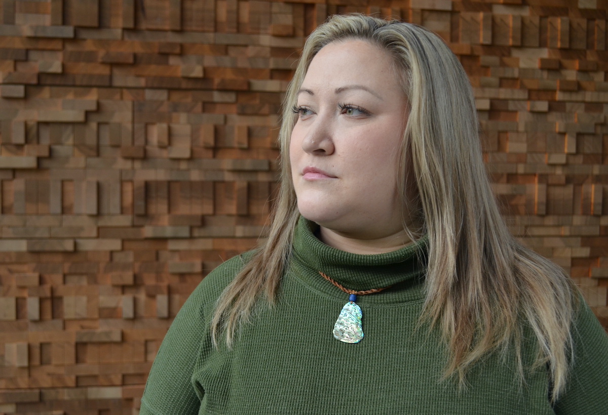 A woman with a green shirt and abalone necklace looks to the left.