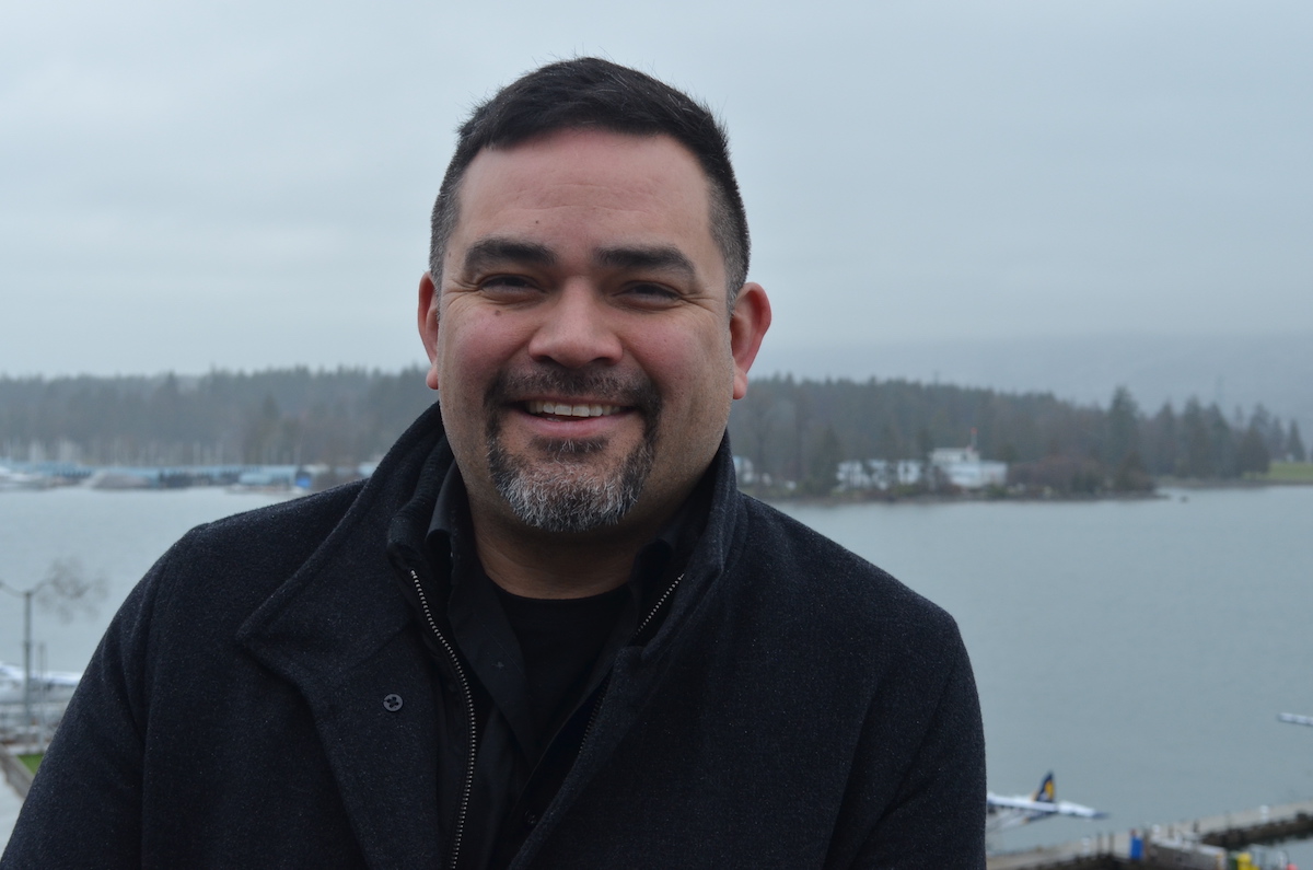 A man in a black jacket grins at the camera. The day is grey and rainy behind him.