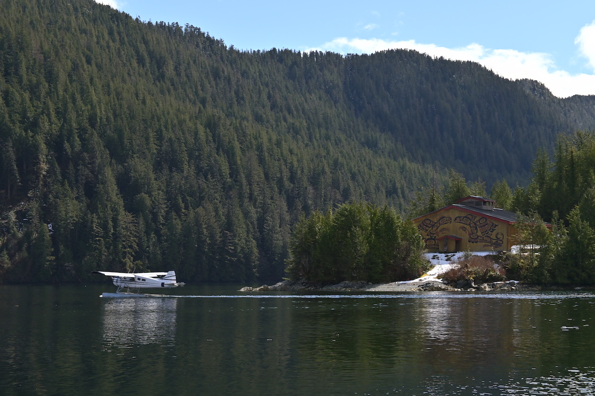 A floatplane lands on the water in front of the shoreline, which is home to a longhouse. 