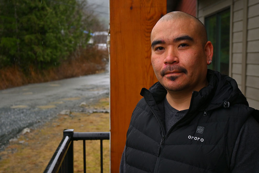 A bald man wearing a black vest looks at the camera. Behind him is a rainy gravel road and green trees.