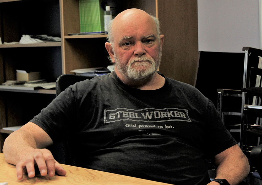 Pat McGregor sits in an office chair looking at the camra and wearing a black T-shirt that says &ldquo;STEELWORKER. And proud to be.&rdquo;