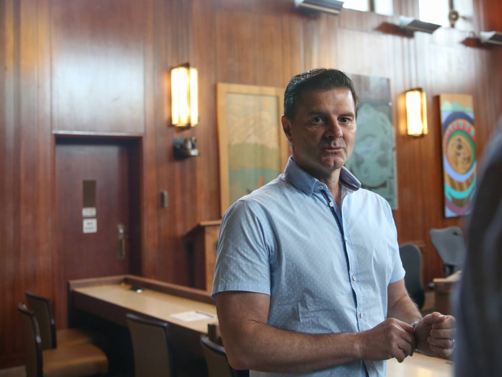 A man in a dress shirt is scrummed by reporters in a council chamber.