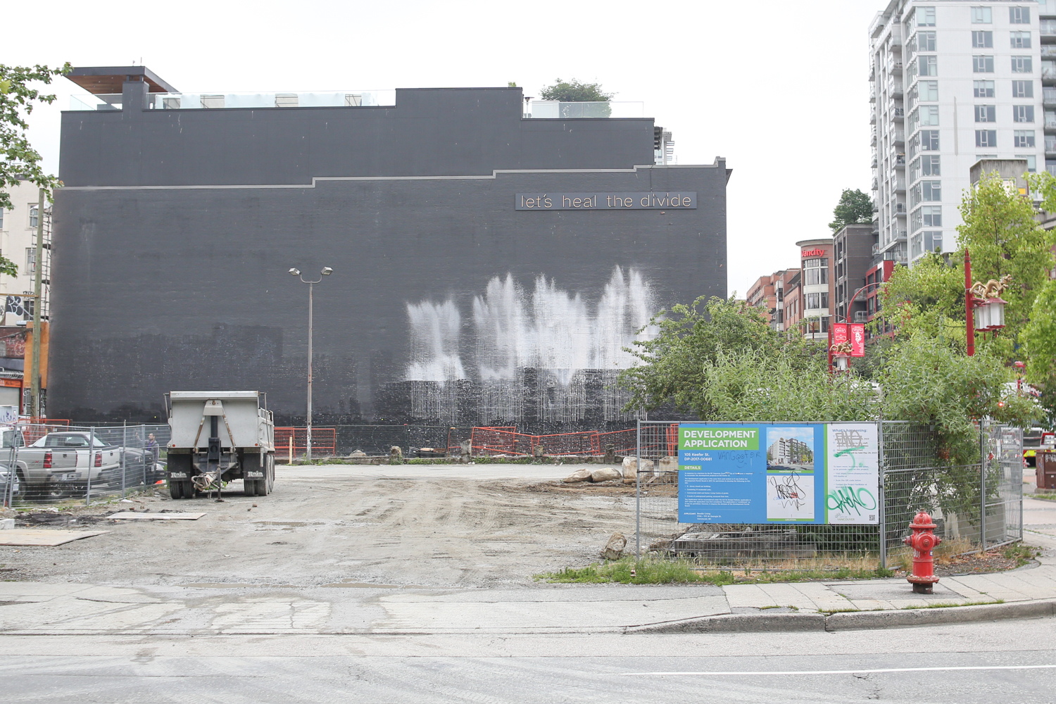 An empty piece of land with some dirt trucks. It is fenced off. On the fence is a bright blue design for a development application with a picture of a condo building on it. On the side of an adjacent building in the back are words in neon: &ldquo;Let&rsquo;s heal the divide.&rdquo;