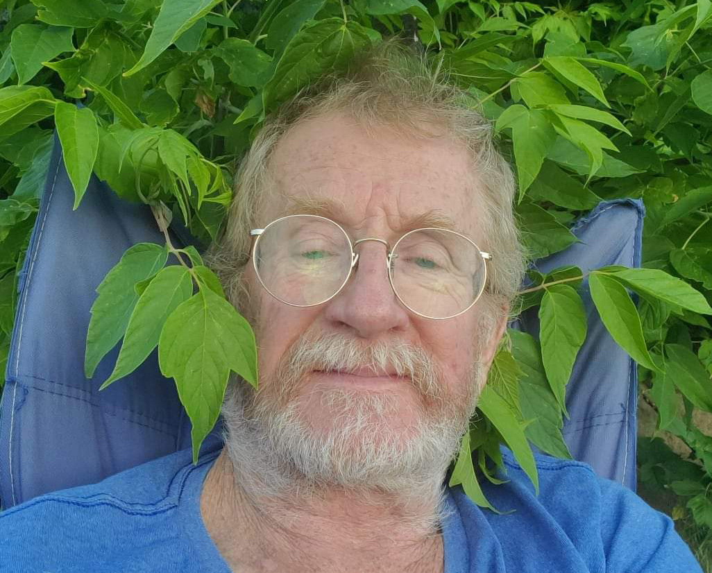 A close-up of a man&rsquo;s face, with greying hair and beard, and wearing glasses. He sits in a blue lawn chair surrounded by green leaves.