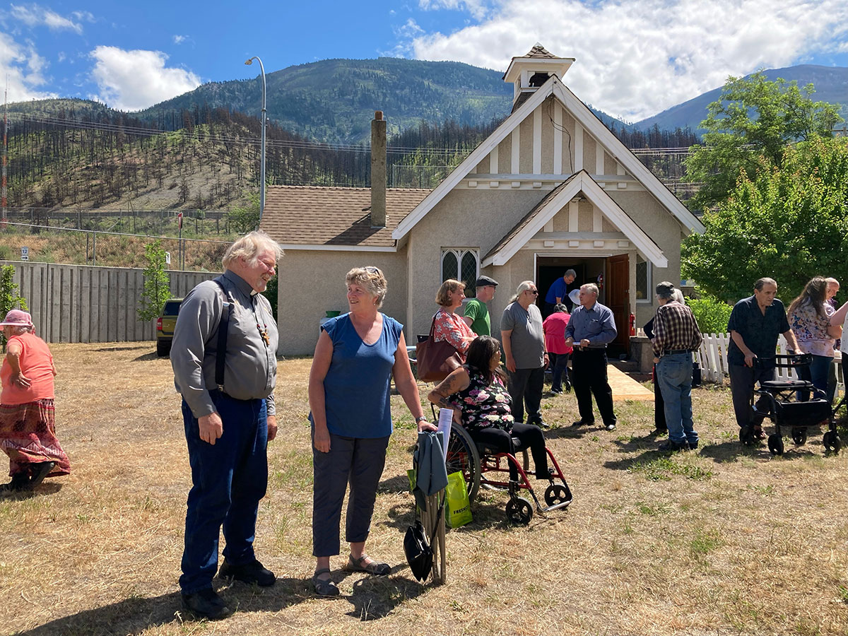 People mingle in front of a small beige building with pointed roof and steeple. On the left, a man with white hair speaks to a woman wearing a blue shirt.