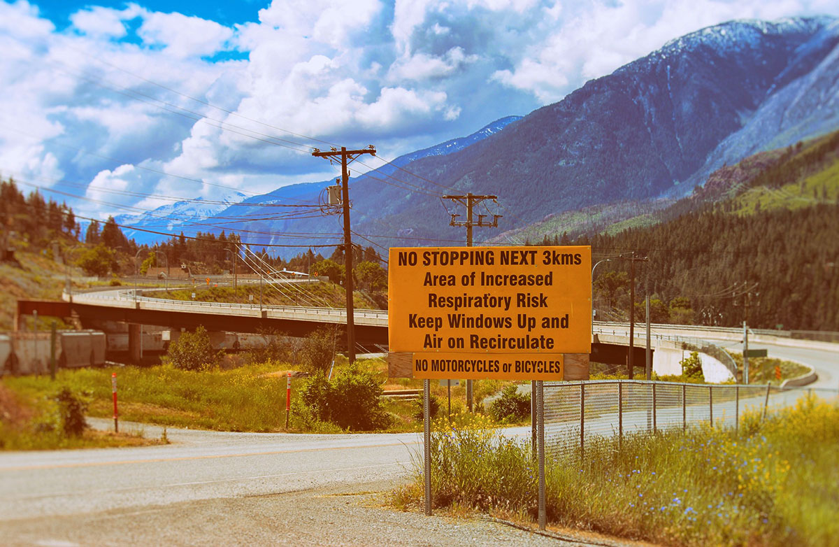 An orange sign next to a road says &ldquo;no stopping next three kilometres, area of increased respiratory risk, keep windows up and air on recirculate.&rdquo; There is mountainous terrain in the background.