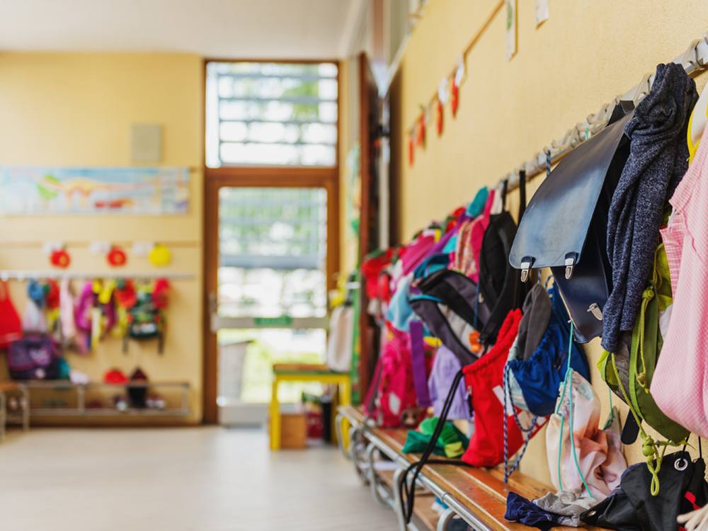 Coat hooks and a bench in a corner of a classroom that opens onto an outdoor setting. There are children&rsquo;s clothes and bookbags hanging on the hooks.