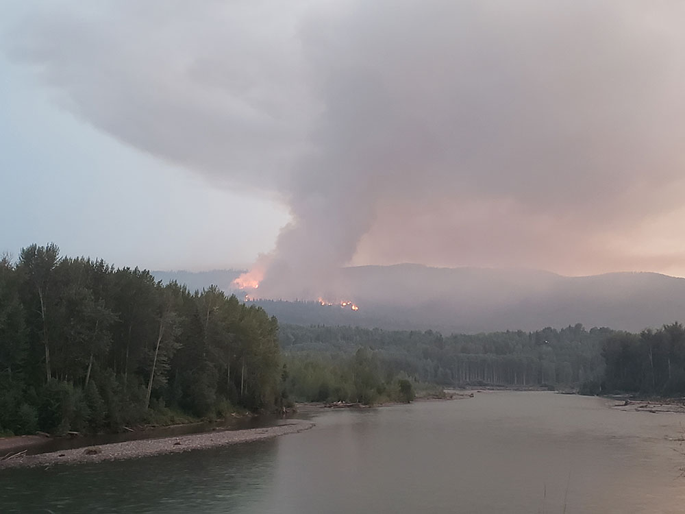 There is a calm body of water in the foreground. Behind, a fire burns on a treed slope, sending a plume of smoke into the air.