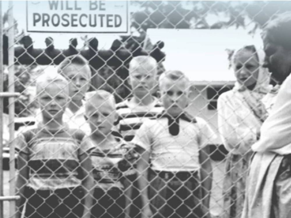 Young boys in striped T-shirts look without emotion through a chain-link fence, while two women look on. A partially obscured sign in the background says 'Will Be Prosecuted.'