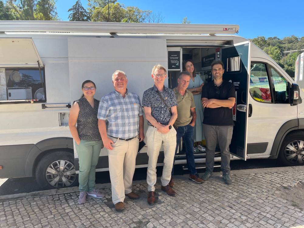 Six people dressed in light-coloured clothes pose in front of a white van. They&rsquo;re smiling and standing casually, with their hands in their pockets or crossed in front of them. 
