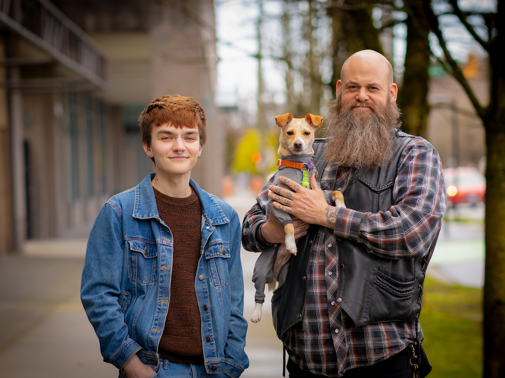 Two people, one of whom is holding a small dog in their arms, stand side-by-side on a sidewalk.