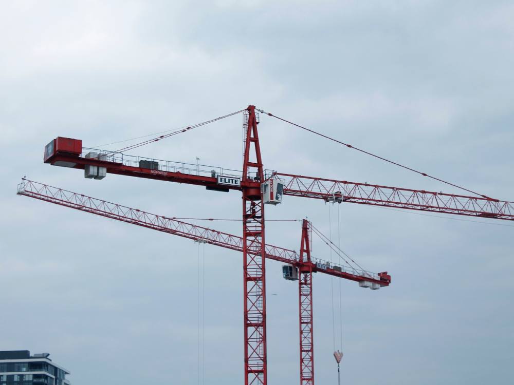 Two red cranes tower above the ground under a cloudy sky.