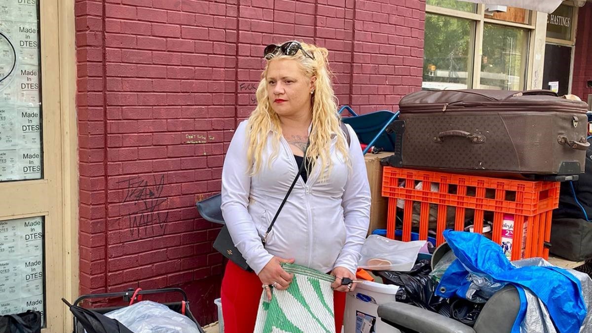 A woman in a white zip-up hoodie with long, wavy blond hair stands surrounded by her belongings in the Downtown Eastside. She is holding a green and white woven mat.