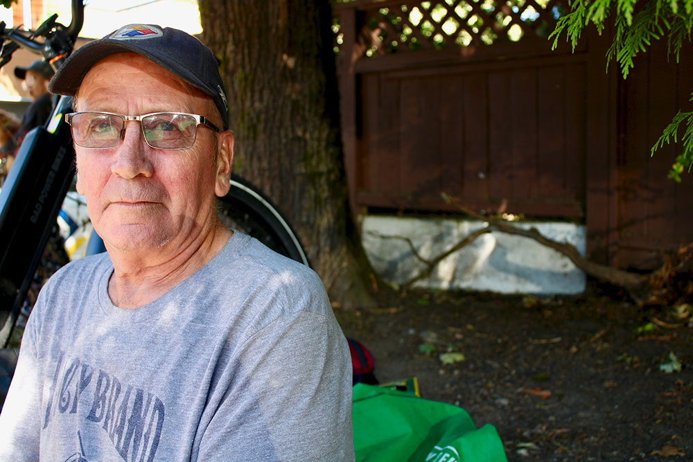 A man in a grey shit, black baseball cap, and glasses looks ahead in front of a brown fence.