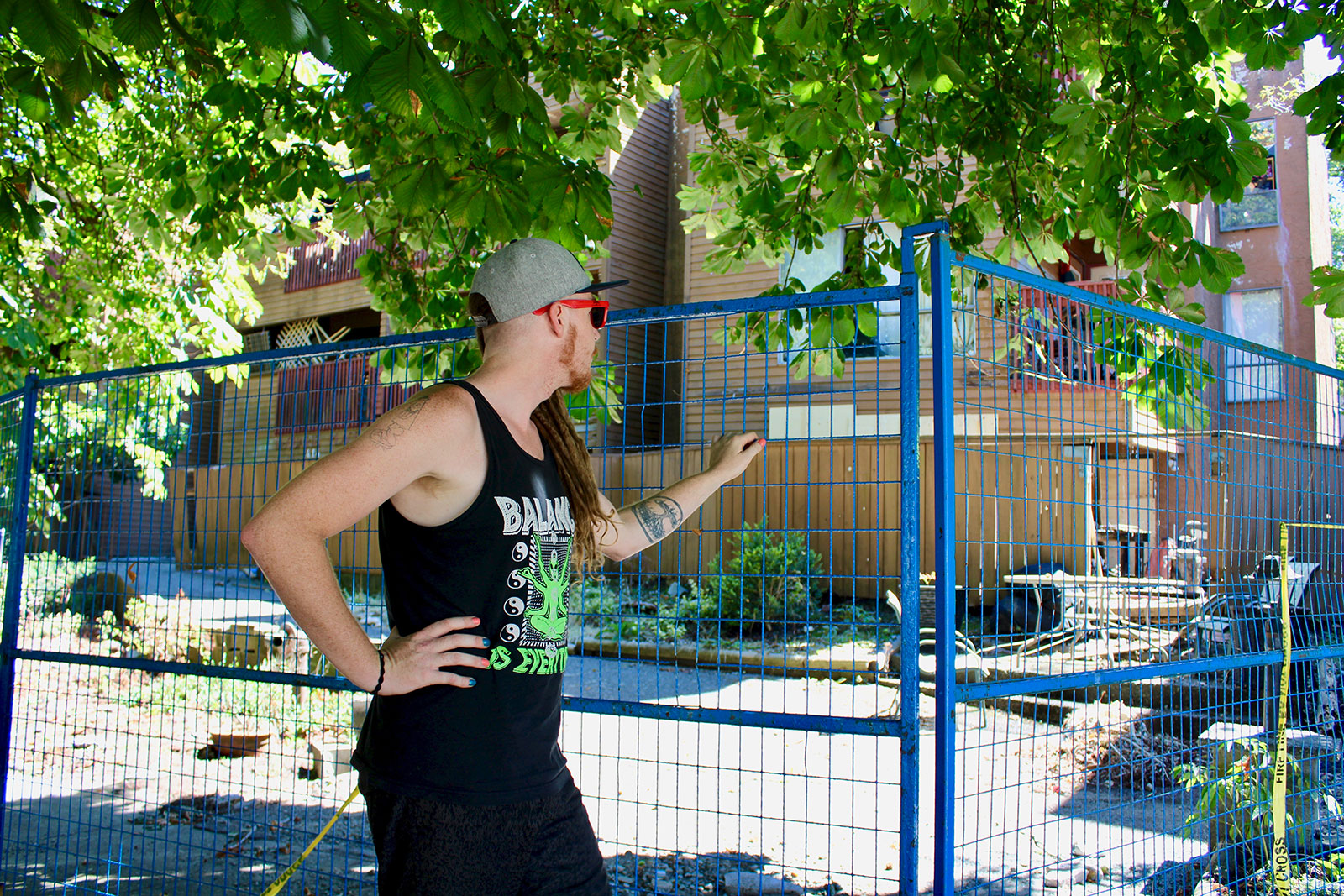A man in a black tank top has his hands on a blue fence, looking at the remains of a scorched building. 