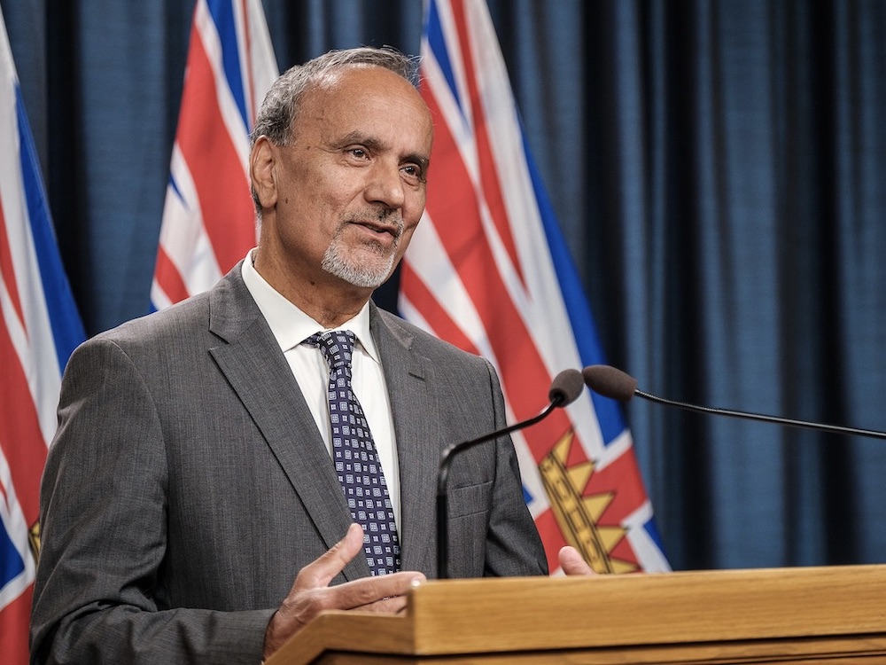 A middle aged man of Indian descent with thinning hair stands at a podium with a microphone, wearing a suit and tie with three British Columbia flags behind him.