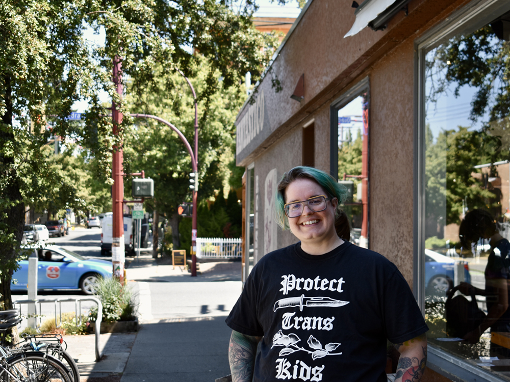 Sloane Chomeakwich stands outside Caffe Fantastico on a sunny day wearing a shirt that says &lsquo;Protect Trans Kids.&rsquo;