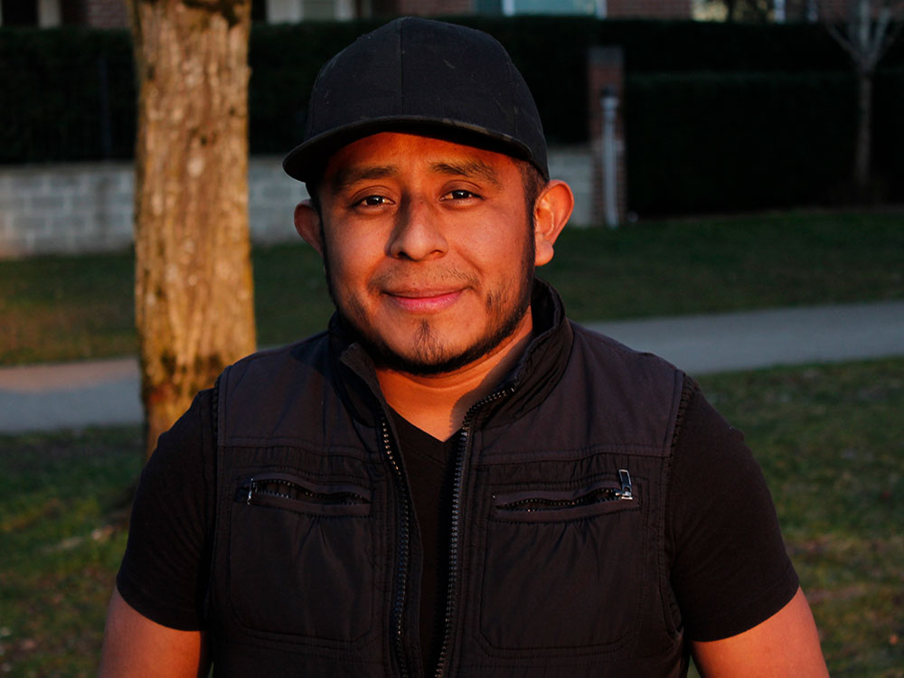 A man in a black shirt, black vest and black ball cap looks steadily at the camera.