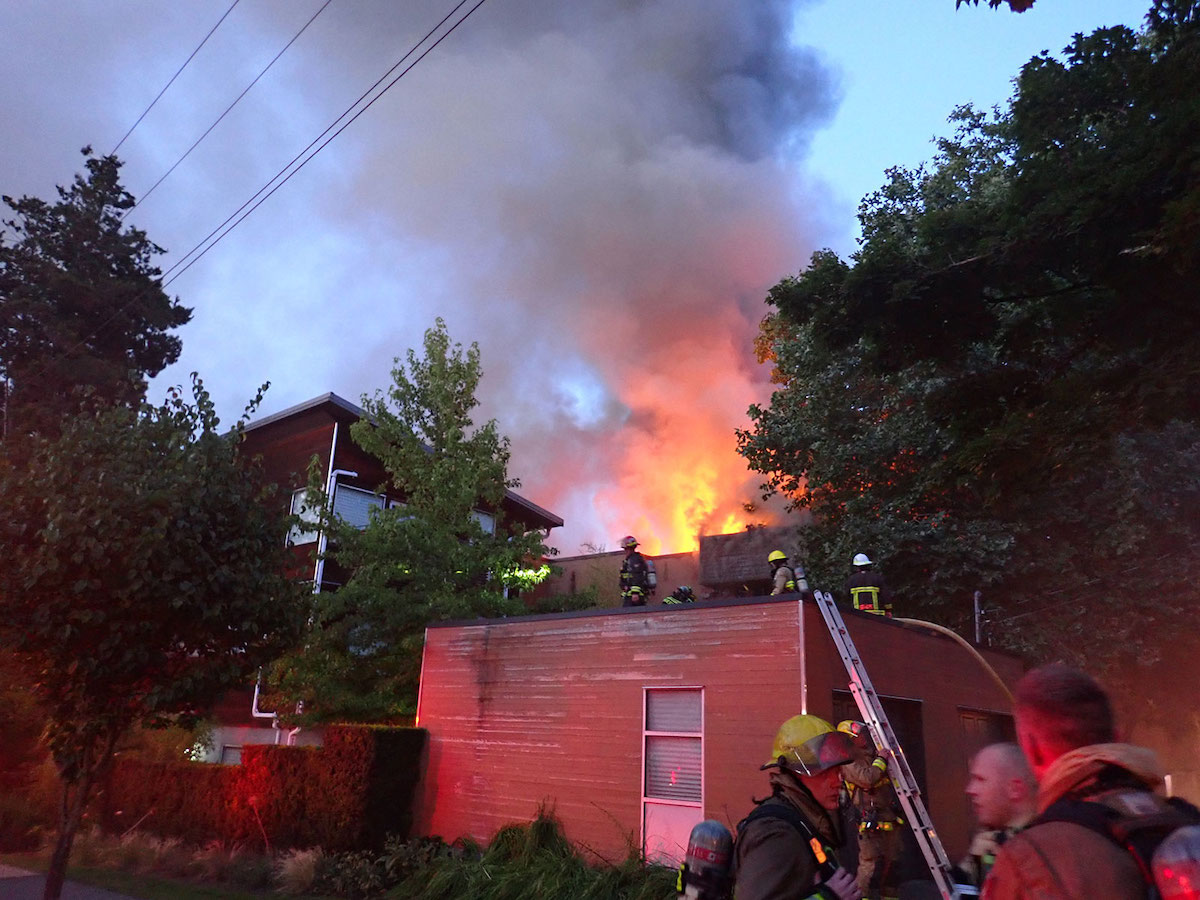 Smoke and fire rise from a low residential building. Firefighters can be seen on the roof of a low building in front of the fire.