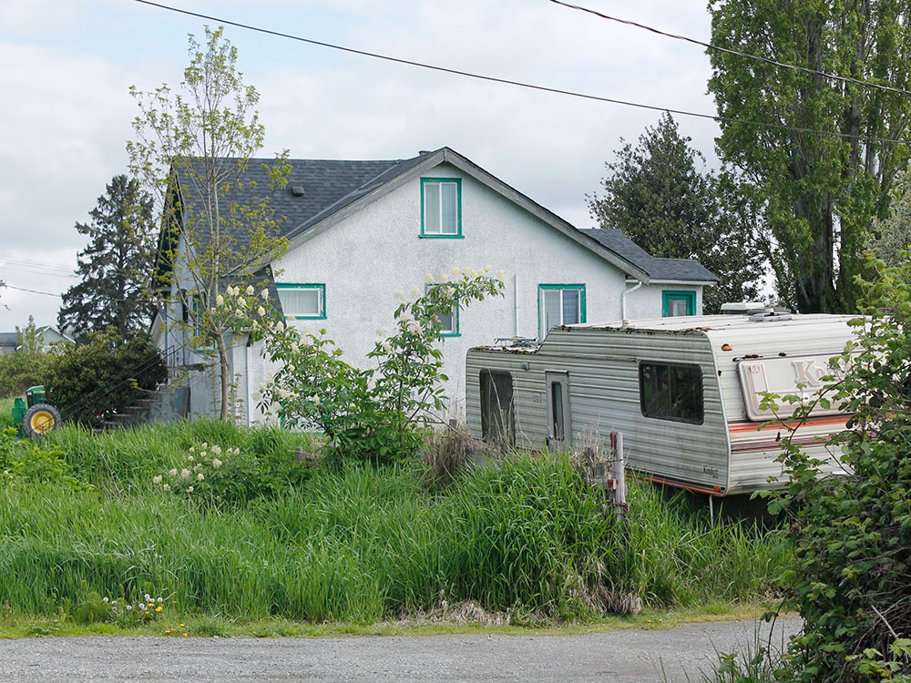 A white stucco house with an RV parked out front. The grass is overgrown.