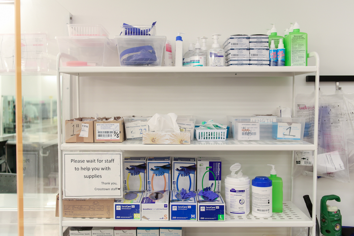 Harm reduction supplies, such as latex gloves, are arranged on a shelving unit. A sign says, 'Please wait for staff to help you with supplies.'