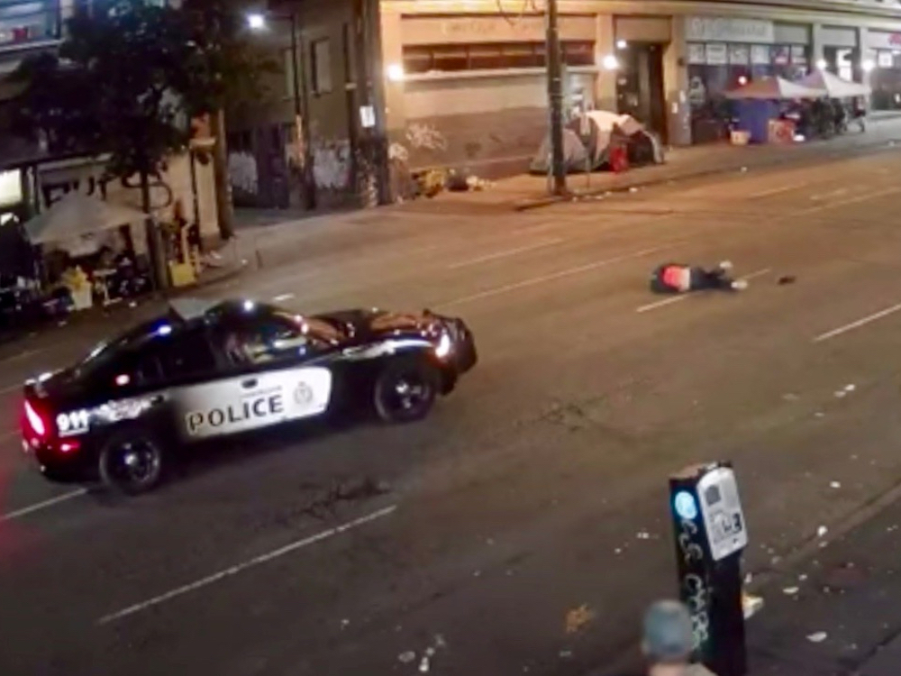 A police car is stopped on East Hastings at night. A body lies sprawled on the road in front of the car.