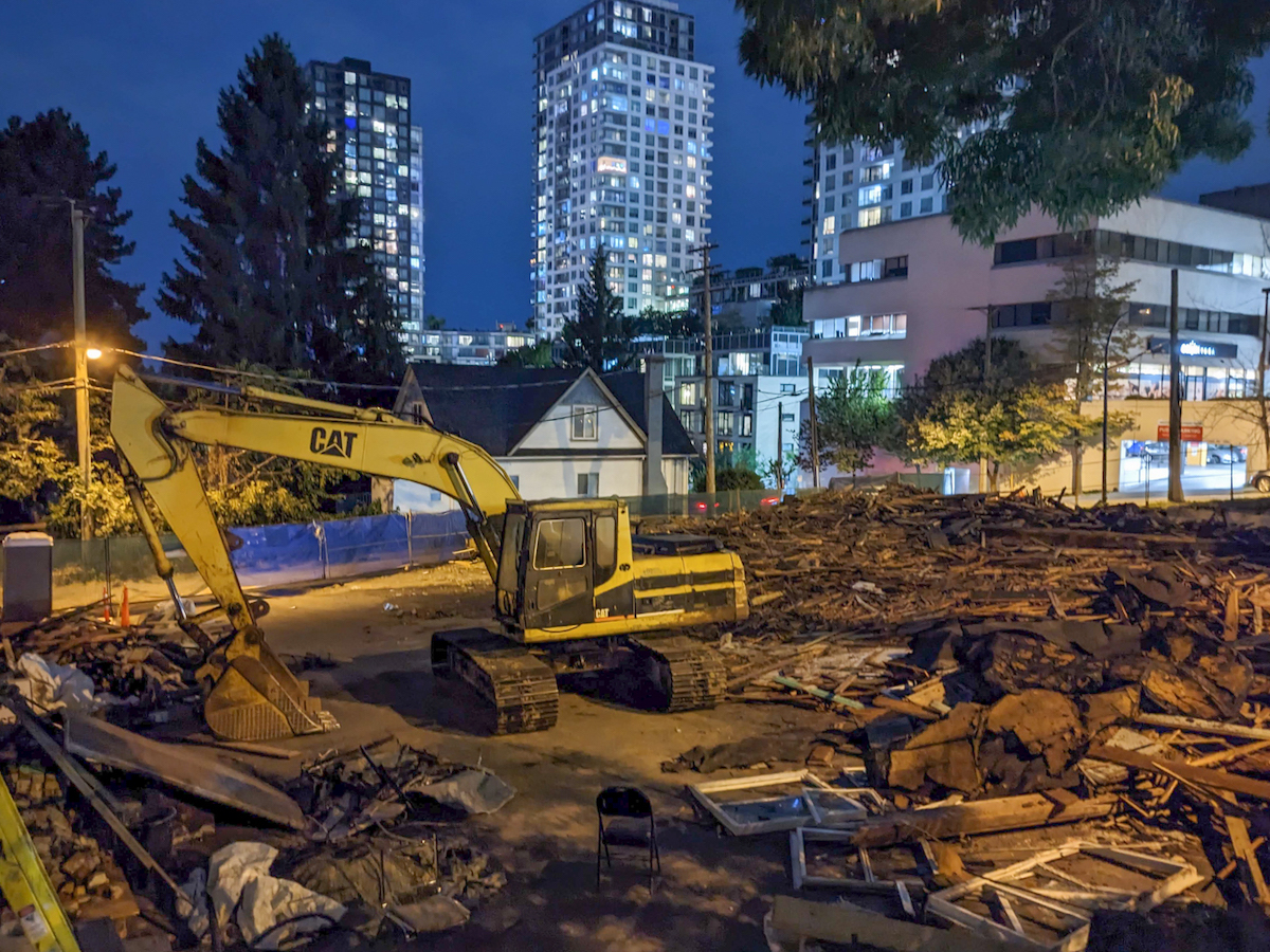 A digger amidst a mess of wood at night. Windows are aglow in the highrises behind.