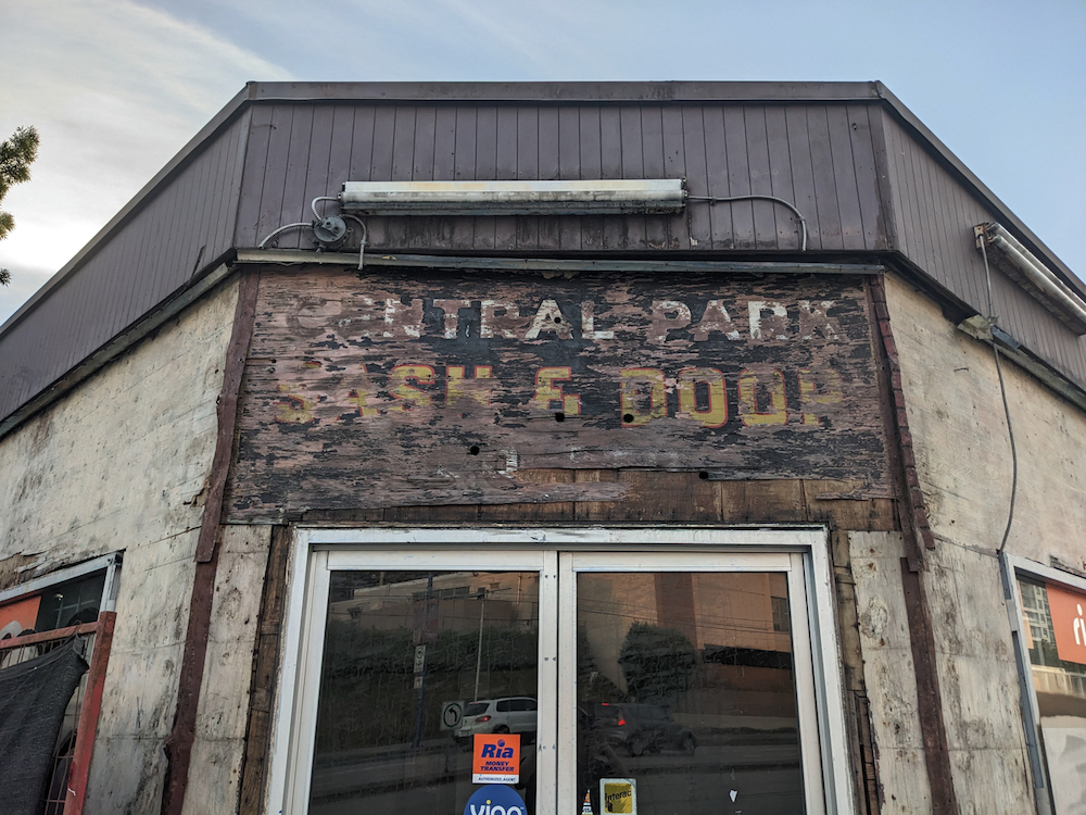 A one-storey building being stripped of its paint, revealing the faded letters of “Central Park Sash & Door” above a modern glass door.