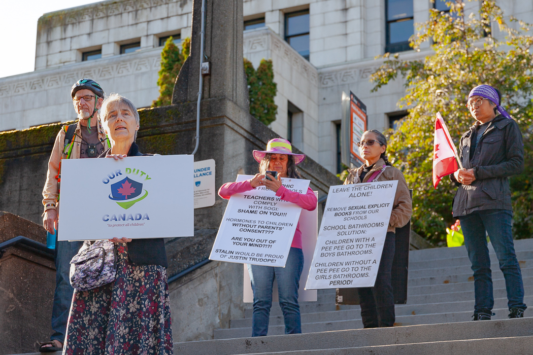 A small group of people gather on the stairs at Vancouver City Hall, many holding signs with transphobic messages on them.