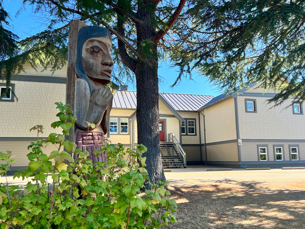 A figure has been carved into a large wooden pole. The figure has their hands together. Their hair is painted black. In the background is a yellow school building with a red door.