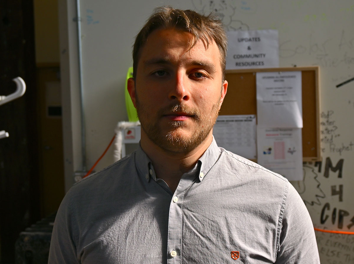 A man in a collared shirt looks at the camera. Behind him is a community notice board and a wall that has been decorated with messages in black marker.