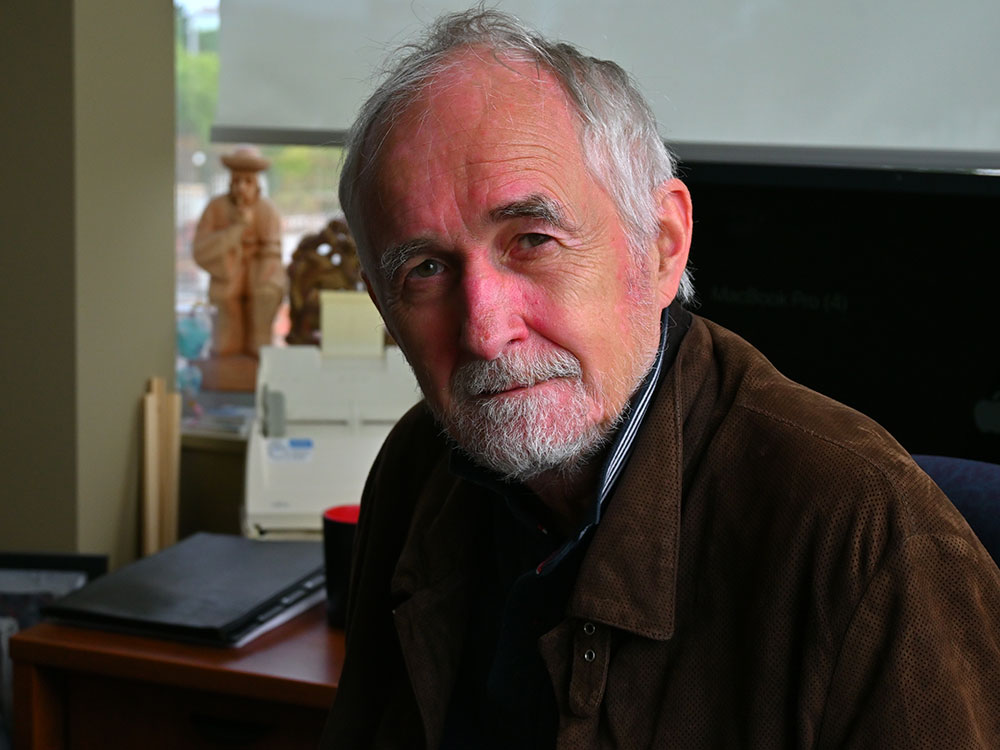 A three-quarters profile of a man with grey hair and a grey beard sitting in front of his computer.