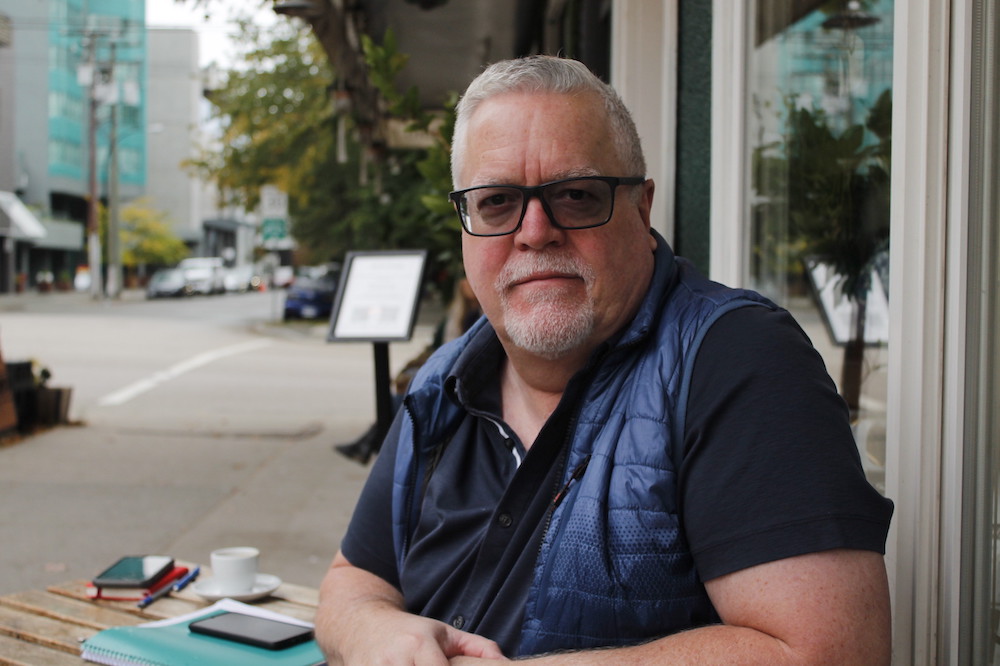 A man wearing a blue shirt and blue vest, with short grey hair and beard and glasses, sits at a café table outside on a grey day.