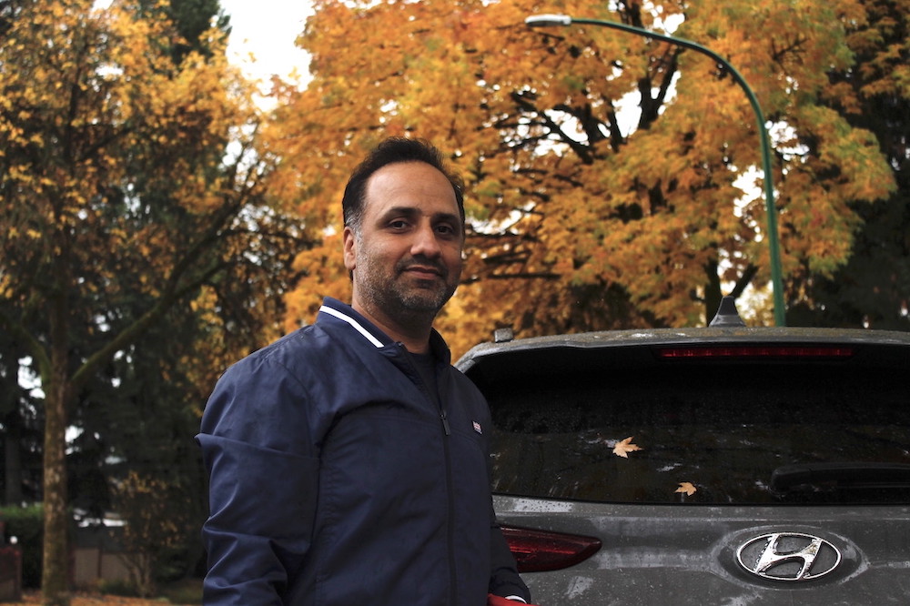 A man wearing a neat blue jacket looks at the camera. He has short dark hair and salt-and-pepper stubble. He is standing in front of a grey car. In the background are trees in their fall colours.