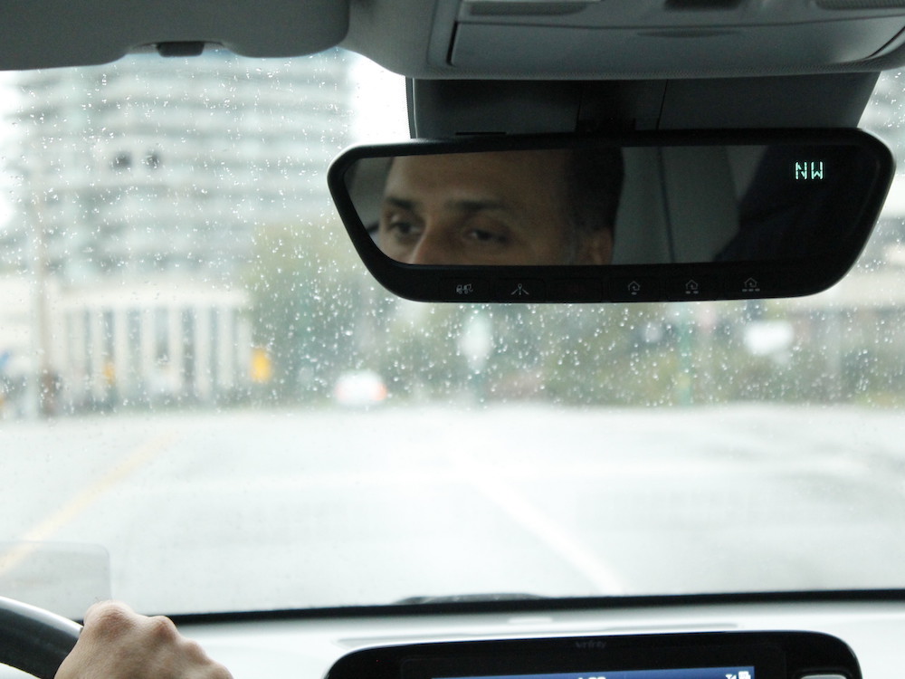 The photo is shot inside the car. One hand grips the steering wheel and we look through the rainy front window. Part of a man's face can be seen in the rear-view mirror.