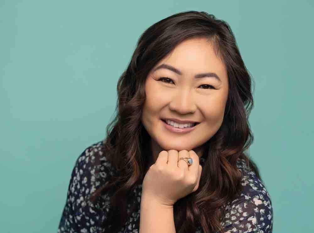 Becca Yu is seated against a turquoise studio background. She has wavy black hair and is wearing a blue and turquoise floral short with dark denim jeans. She is leaning forward slightly with her chin resting against her left hand, smiling.