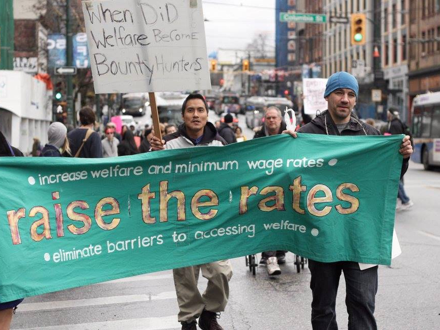 Two people carry a green banner that says 'Raise the Rates' on a Vancouver street under grey skies.