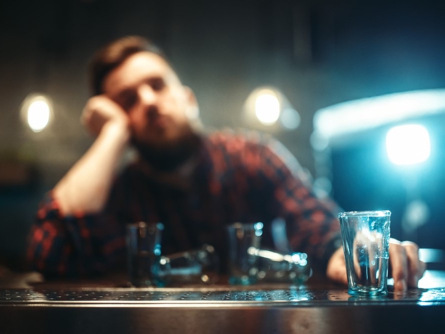 A blurred photo of a person in a black and red plaid shirt sitting on the far side of a bar. An empty glass is in the foreground.