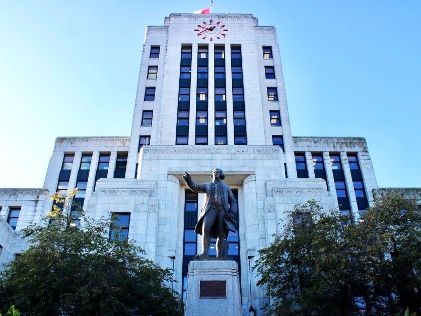 The grey facade of Vancouver City Hall looms above a statue of a man standing on a plinth and gesturing. The sky is pale blue.