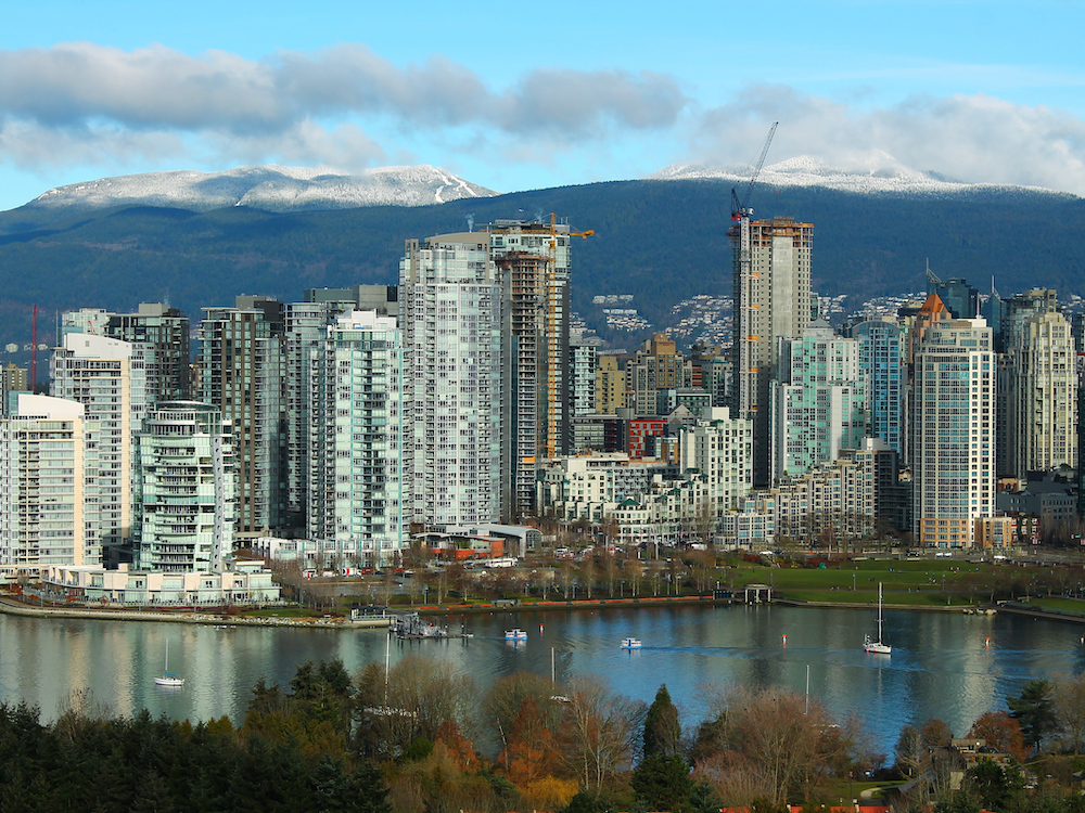 The skyline of Vancouver shows water, tall buildings and snow-dusted mountains under a blue sky.