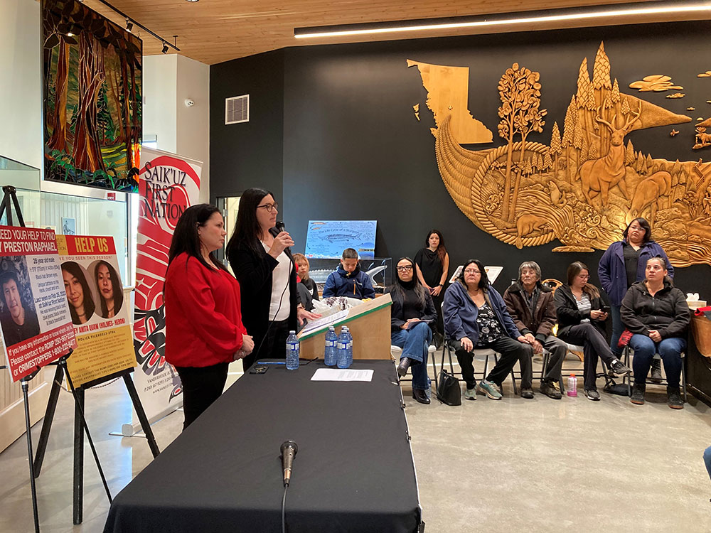 At left, two women stand with a microphone in front of two missing-persons posters. Several people sit in chairs listening.