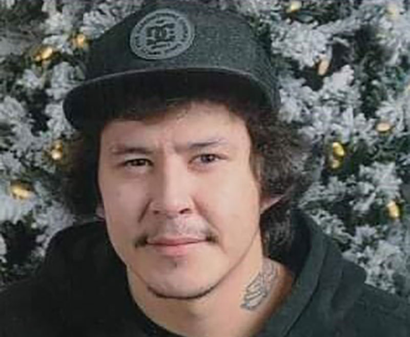 A man wearing a black ballcap and black hoody looks at the camera. He has dark hair and mustache and a tattoo on his neck.