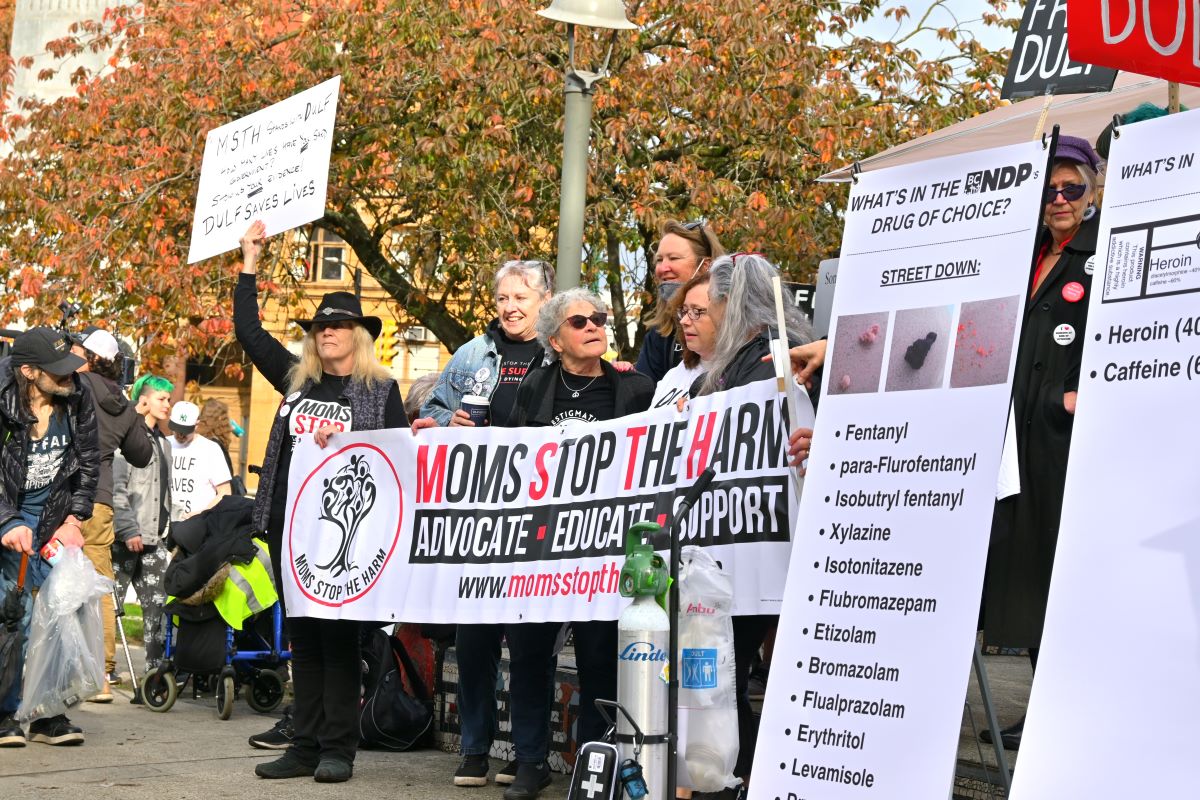 Moms Stop the Harm at a rally for DULF. A group of women holds a banner that says, &lsquo;MOMS STOP THE HARM.&rsquo; Another person holds a large portrait-orientation sign that lists many substances that have been mixed into street drugs. 