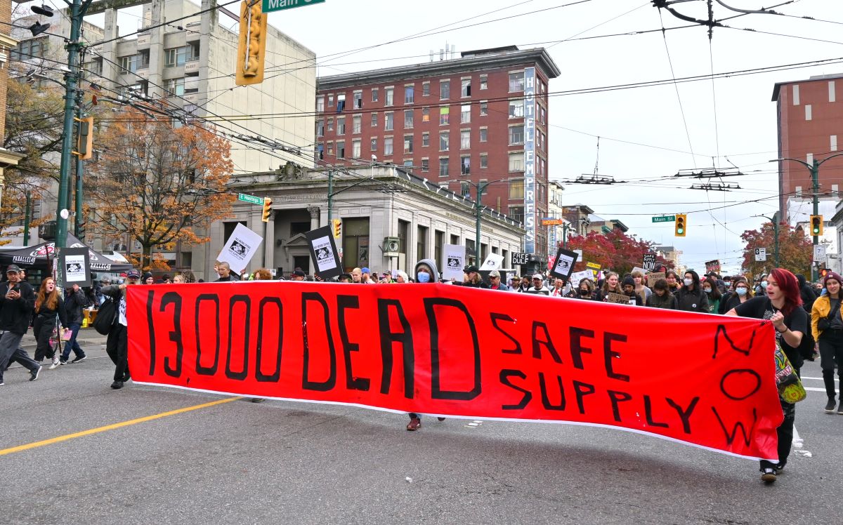 The rally for DULF moves through the downtown eastside. A group of people hold a banner that reads, &lsquo;13,000 DEAD. SAFE SUPPLY NOW.&rsquo;