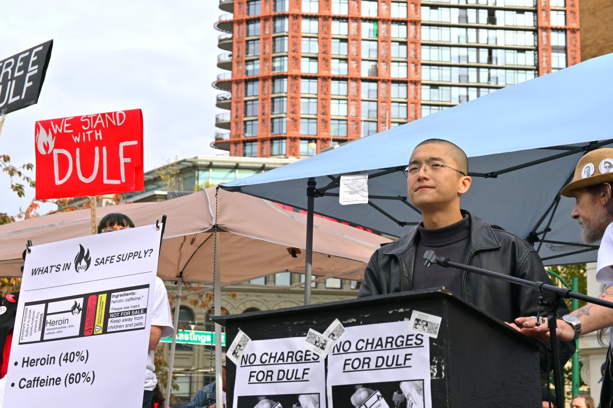 Vince Tao, a man with a shaved head and olive skin tone, stands at a podium at Victory Square in Vancouver. Posters taped to the podium read, &lsquo;No Charges for DULF.&rsquo;