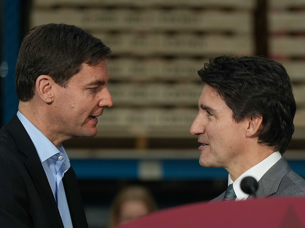 A white man with short black hair, a dark suit and blue shirt stands facing another white man with longer hair, a grey suit, white shirt and blue tie.