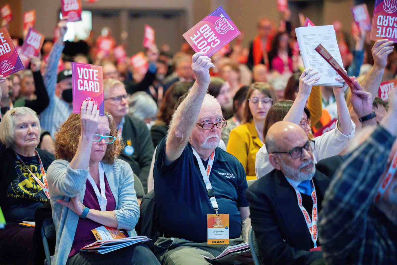 A large group of people sitting in rows of folding chairs hold up red voting cards.